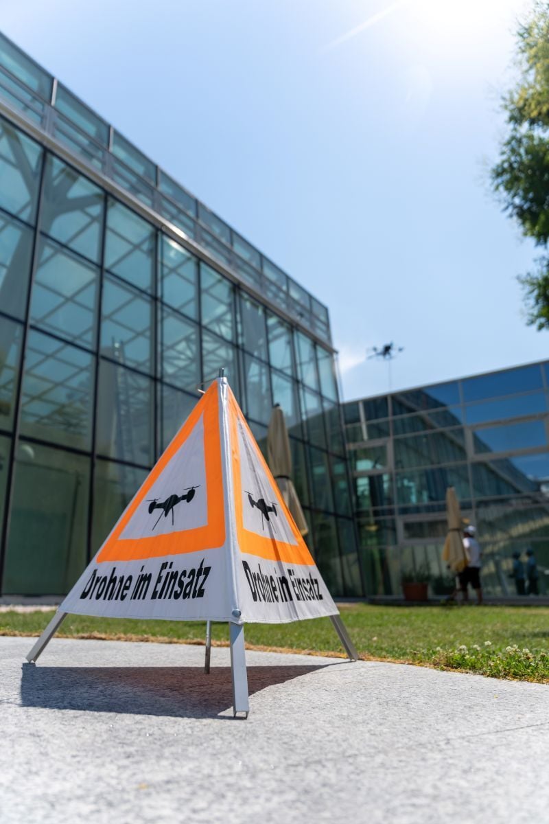 Warnschild „Drohne im Einsatz“ vor einem modernen Bürogebäude, während im Hintergrund eine Drohne Reinigungsarbeiten durchführt.