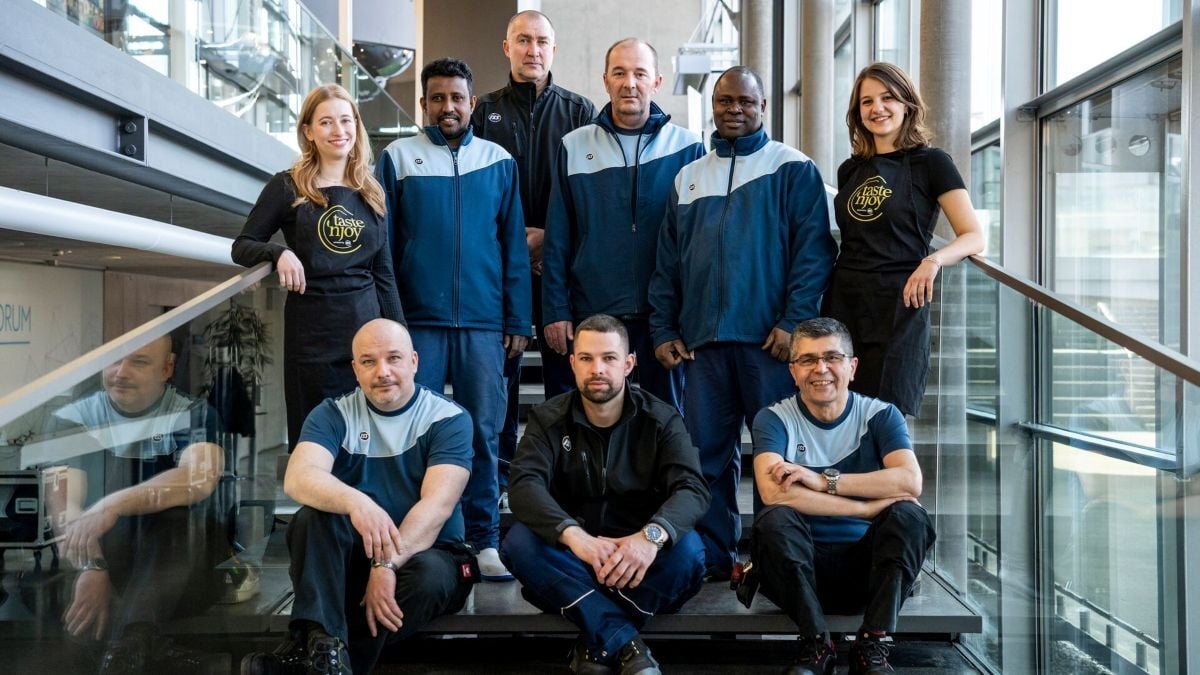 Gruppe von Personen in ISS‑Arbeitskleidung und „taste ’n joy“-Schürzen steht und sitzt auf einer Treppe in einem modernen Gebäude mit Glasgeländern