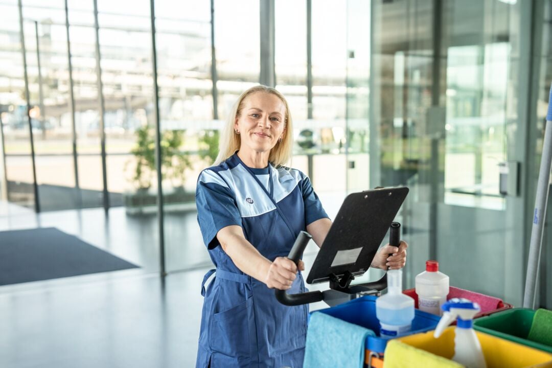 ISS Reinigungskraft in Arbeitskleidung steht hinter einem Reinigungswagen mit Tablet und Reinigungsmitteln in einem modernen Gebäudeinnenbereich