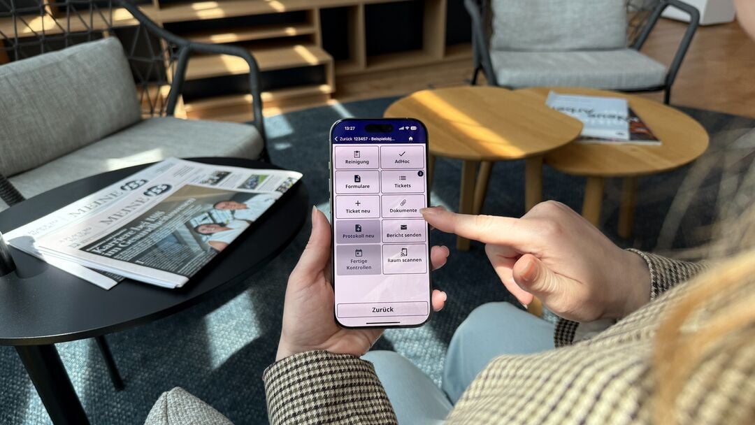 Person hält ein Smartphone mit einer App‑Übersicht für Reinigung und Dokumente und tippt auf den Menüpunkt „Dokumente“, im Hintergrund stehen Sessel und Tische