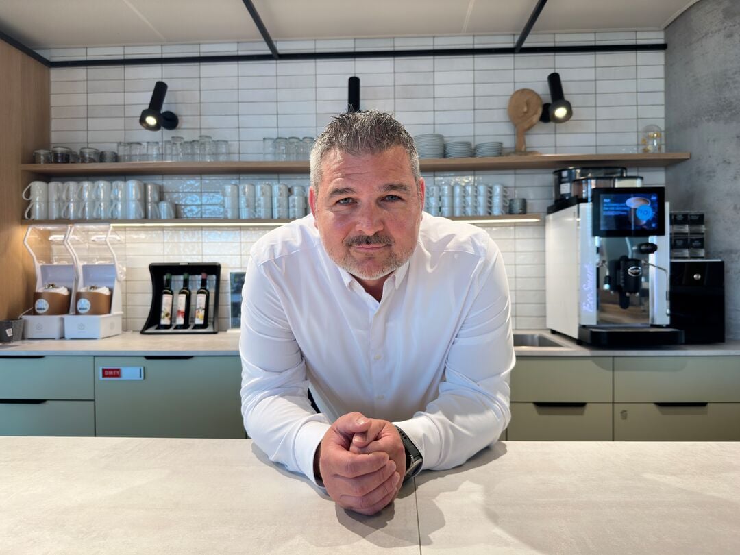 Person in weißem Hemd lehnt an einer Theke in einem gastronomischen Innenbereich mit Kaffeemaschine, Gläsern und Regalen im Hintergrund