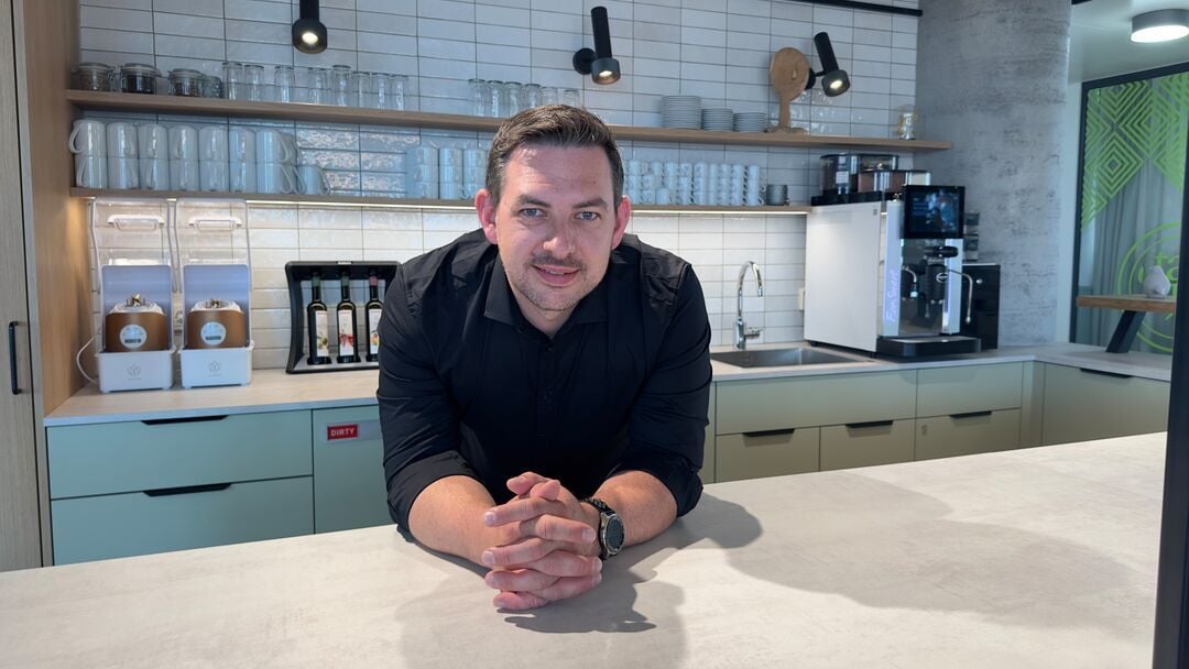 Person in dunklem Hemd lehnt an einer Theke in einem gastronomischen Innenbereich mit Kaffeemaschine, Gläsern und Regalen im Hintergrund