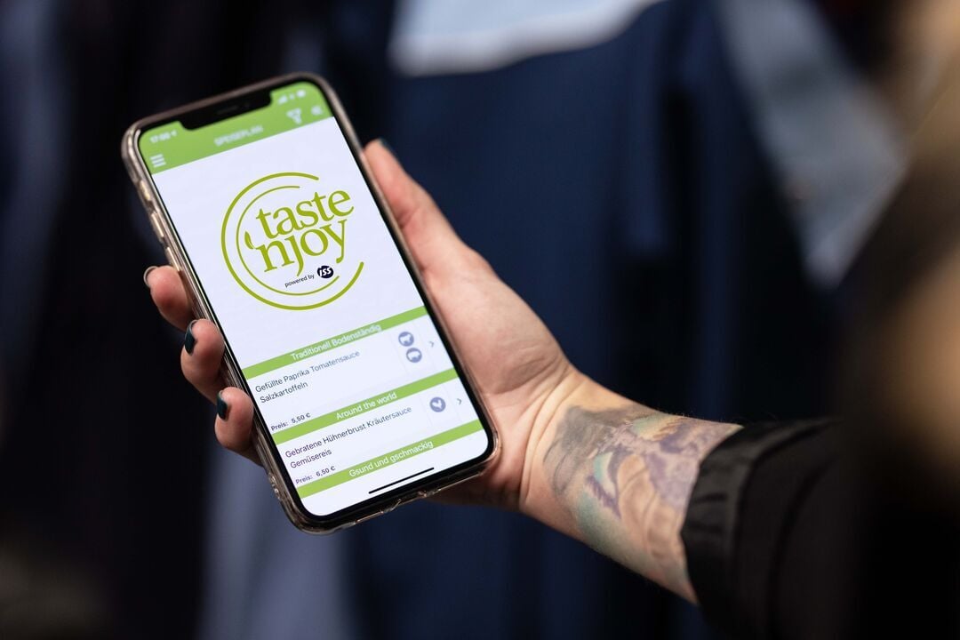 Hand hält ein Smartphone mit geöffneter „taste ’n joy powered by ISS“-App, auf dem Logo und Menüeinträge angezeigt werden