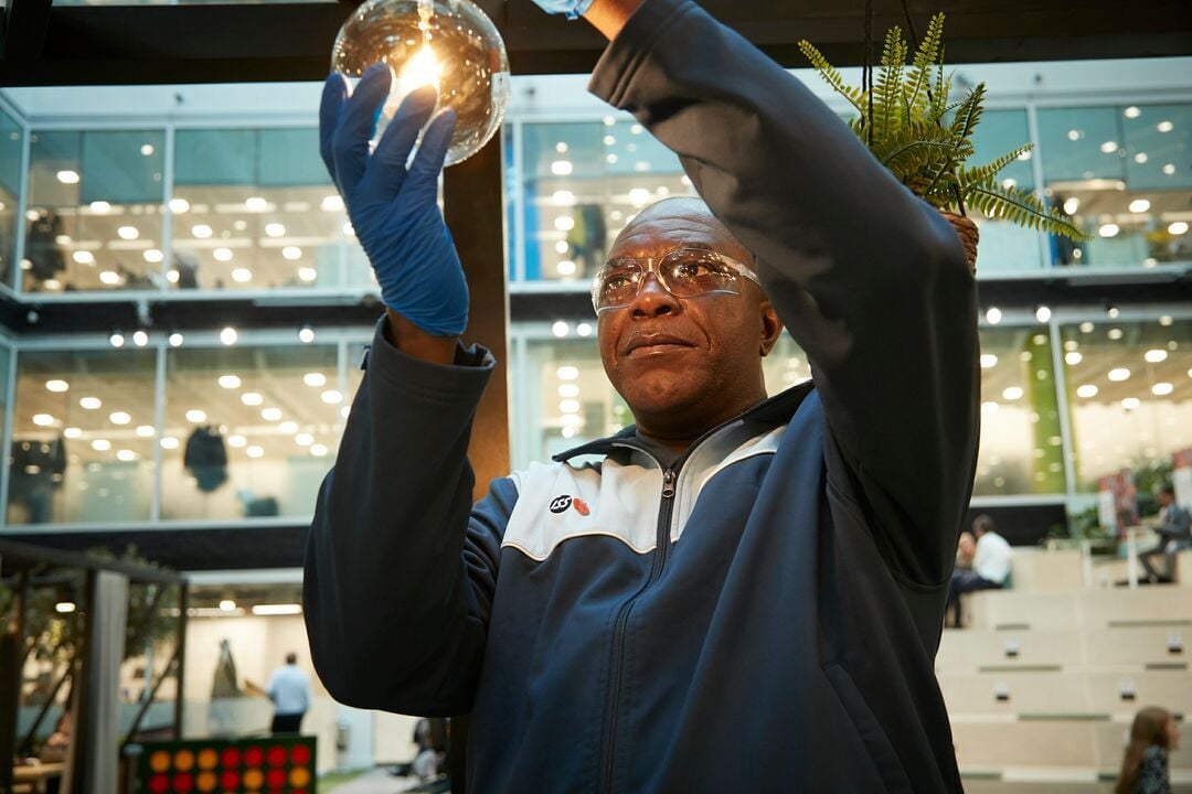 Person in ISS Arbeitskleidung und mit Handschuhen hält eine leuchtende Glühlampe in einem modernen Innenraum mit Sitzstufen und Glasfassade.