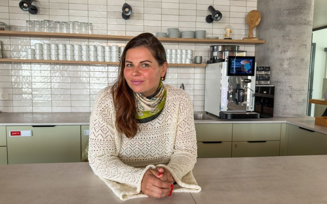 Person mit hellem Strickpullover und Halstuch lehnt an einer Theke in einem gastronomischen Innenbereich mit Kaffeemaschine, Tassen und Regalen im Hintergrund