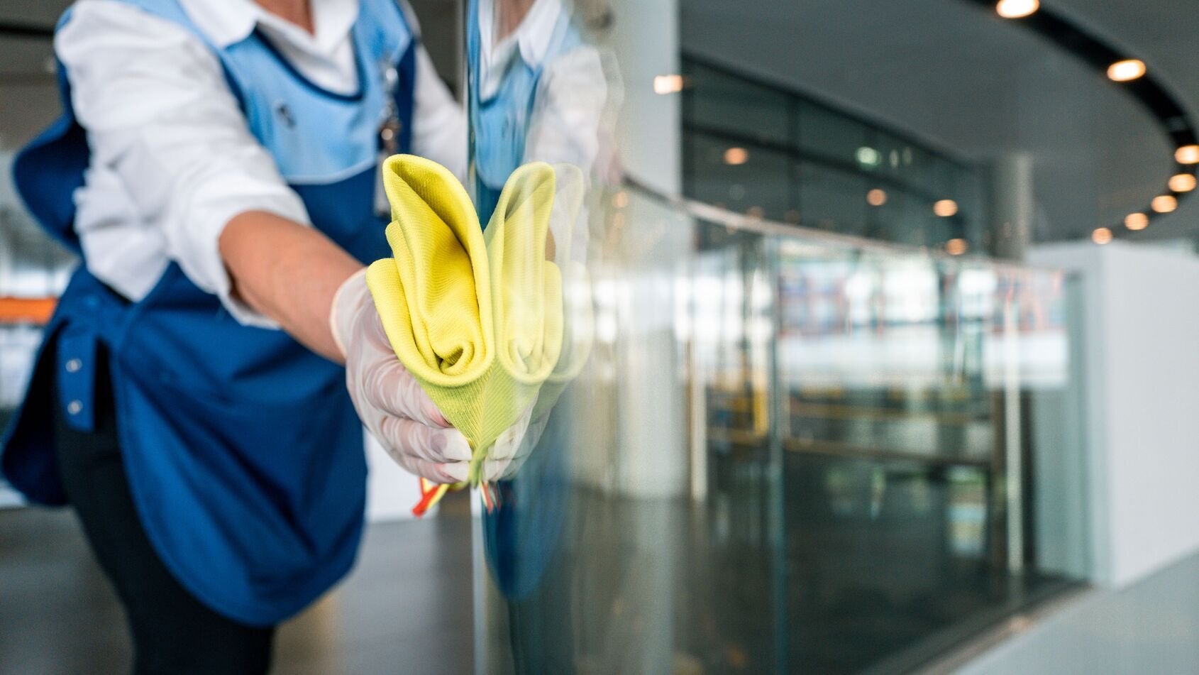 Person mit Handschuhen wischt mit einem gelben Reinigungstuch eine Glasfläche in einem Innenbereich