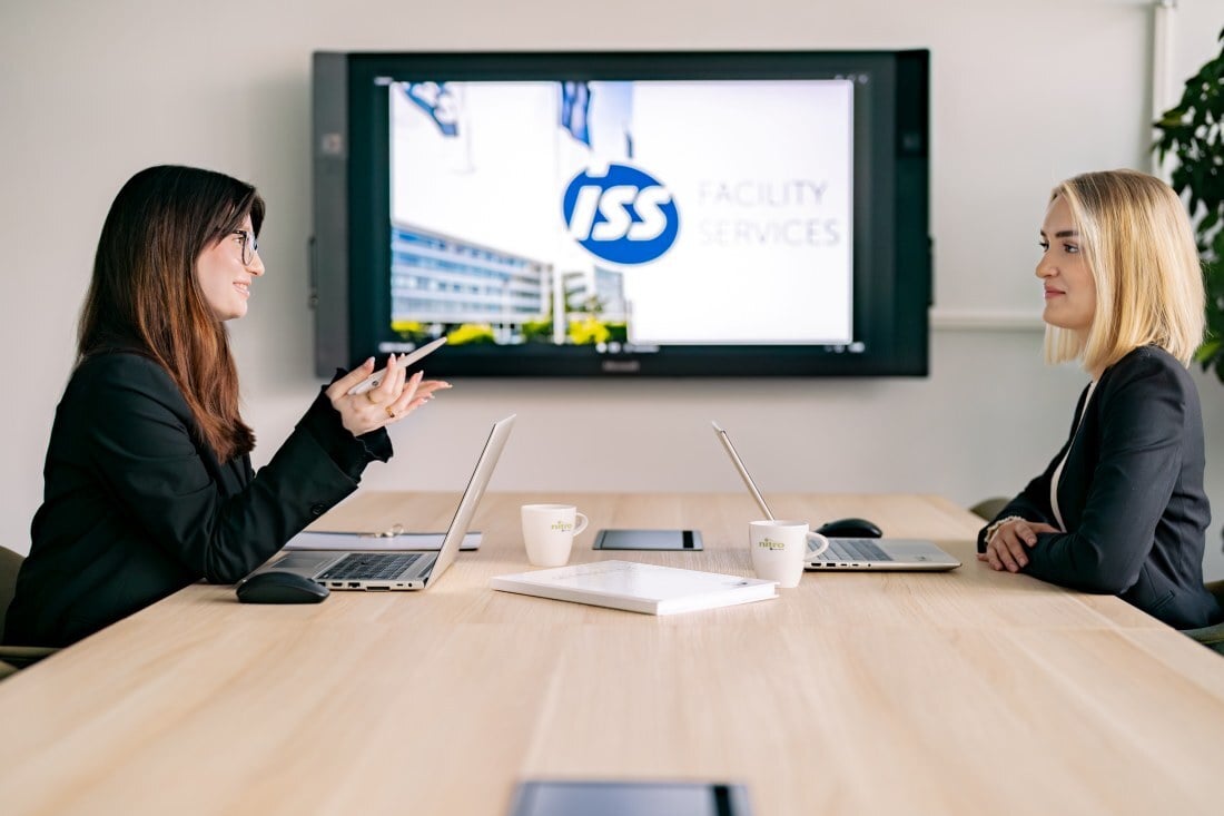Zwei Personen sitzen an einem Tisch mit Laptops in einem Besprechungsraum, im Hintergrund ist ein Bildschirm mit dem ISS‑Facility‑Services‑Logo sichtbar