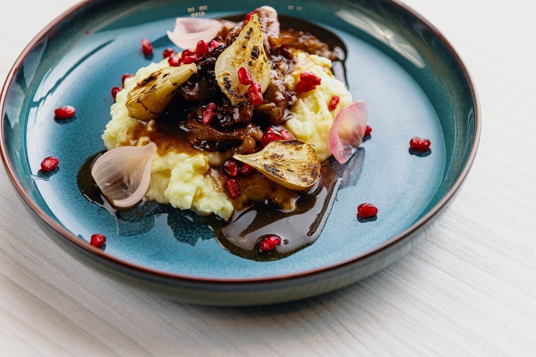 Ansicht von oben auf einen Teller mit Kartoffelpüree, gegartem Fleisch, Karotten, gebratenen Zwiebeln, Granatapfelkernen und dunkler Sauce auf einer hellen Holzoberfläche