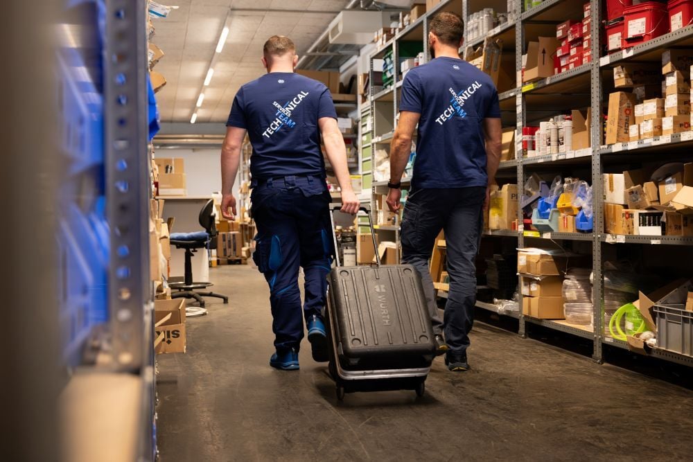 Zwei Mitarbeitende des technischen Service transportieren Werkzeug durch ein Ersatzteillager mit Regalen und Materialbeständen.