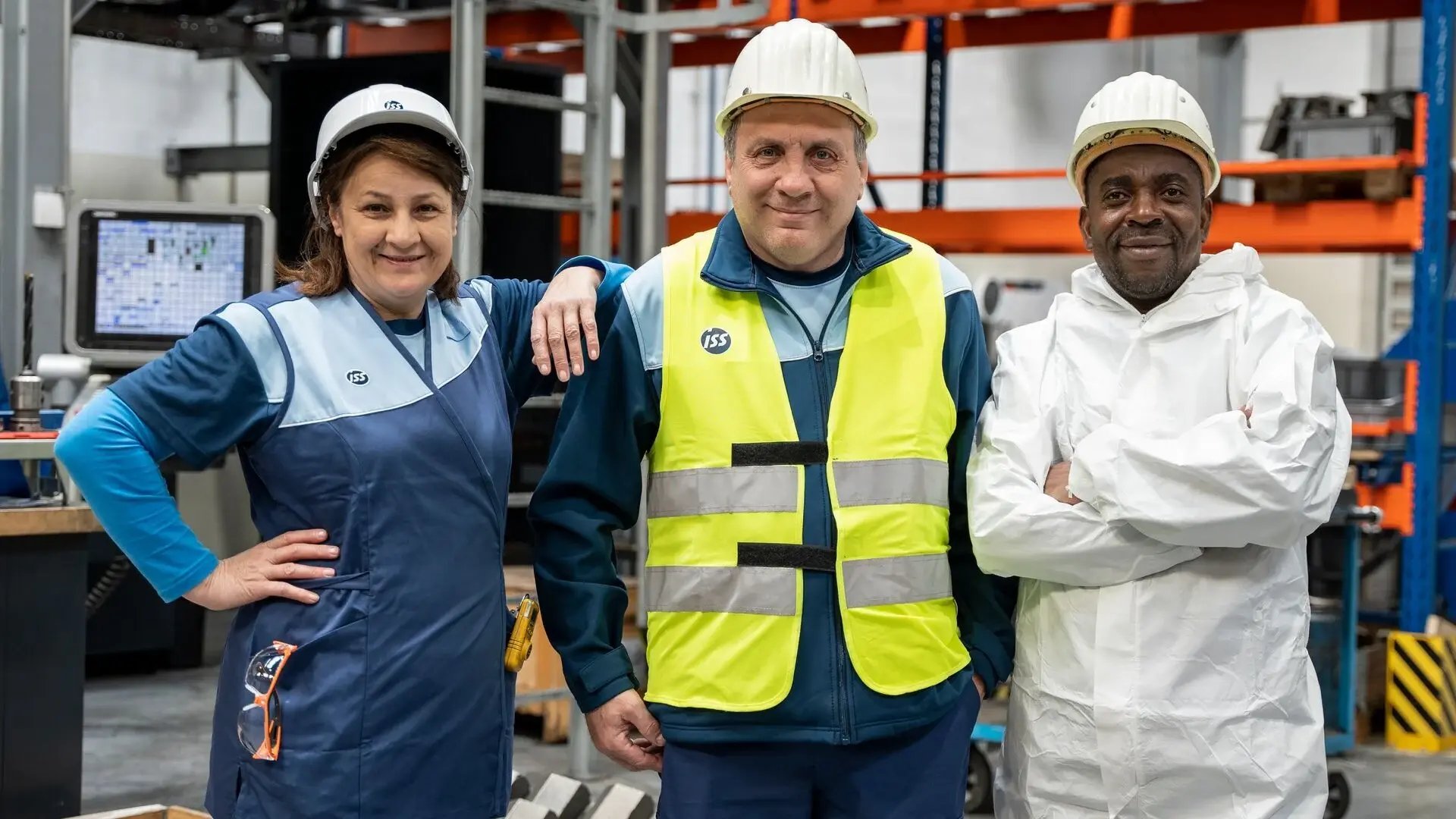 Drei ISS Mitarbeitende in Arbeitskleidung und Schutzausrüstung posieren gemeinsam in einer Produktions- oder Logistikumgebung.