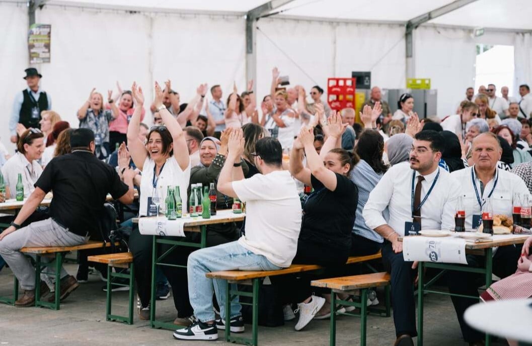 ISS Mitarbeitende sitzen an Bierbänken in einem Festzelt, heben die Arme und reagieren gemeinsam auf das Geschehen; auf den Tischen stehen Getränke.