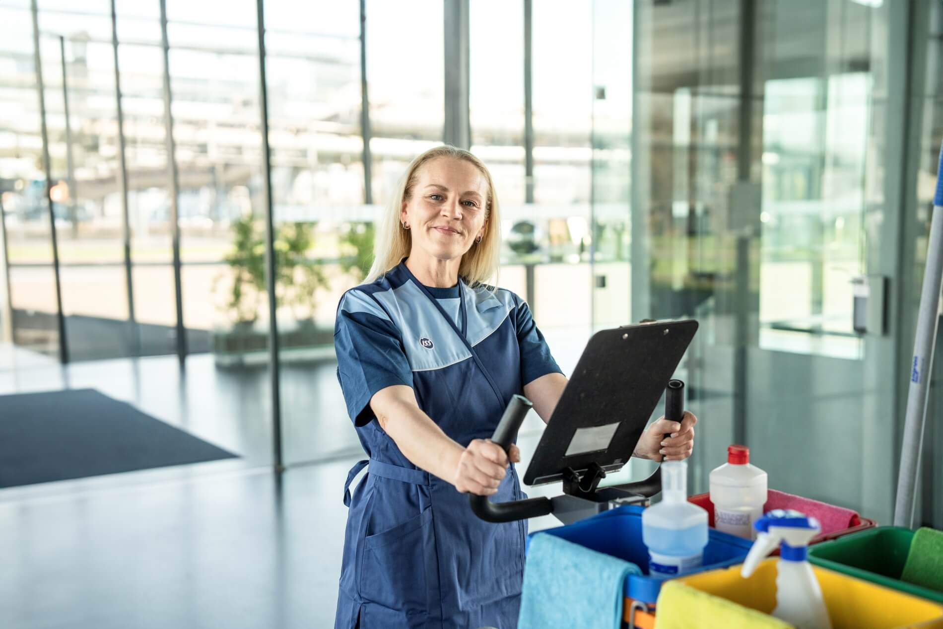 ISS Reinigungskraft in Arbeitskleidung steht hinter einem Reinigungswagen mit Tablet und Reinigungsmitteln in einem modernen Gebäudeinnenbereich