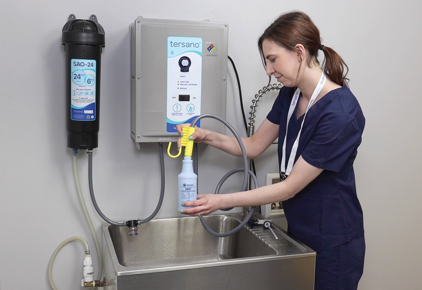 Person in Arbeitskleidung füllt eine Sprühflasche an einem wandmontierten Tersano‑Gerät über einem Edelstahlwaschbecken