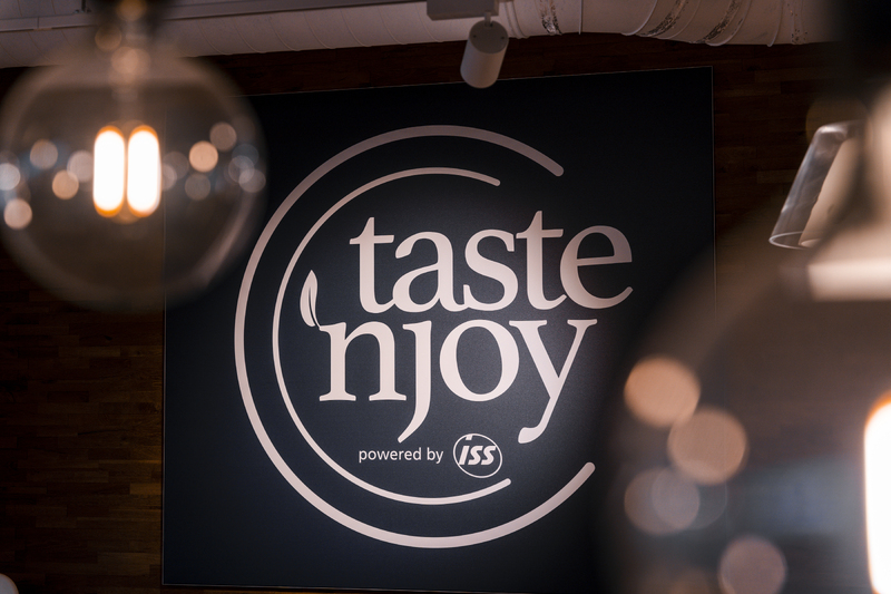 Wandlogo mit der Aufschrift „taste ’n joy powered by ISS“ in einem gastronomischen Innenbereich mit Hängelampen
