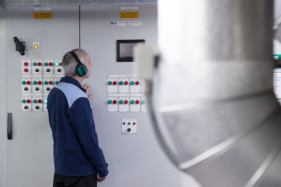 Person mit Gehörschutz steht vor einem elektrischen Schaltschrank mit Tastern und Kontrollleuchten in einem technischen Anlagenraum