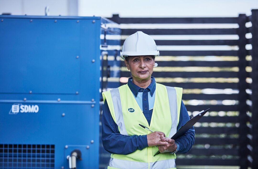 ISS Mitarbeiterin in Arbeitskleidung mit Warnweste und Schutzhelm hält ein Klemmbrett und steht im Außenbereich neben einem technischen Aggregat