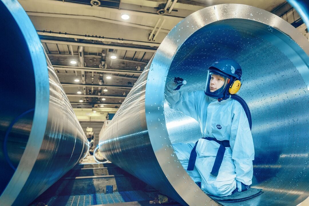 Person in Schutzkleidung mit Helm, Visier und Gehörschutz befindet sich im Inneren eines großen zylindrischen Metallkanals in einer Industriehalle