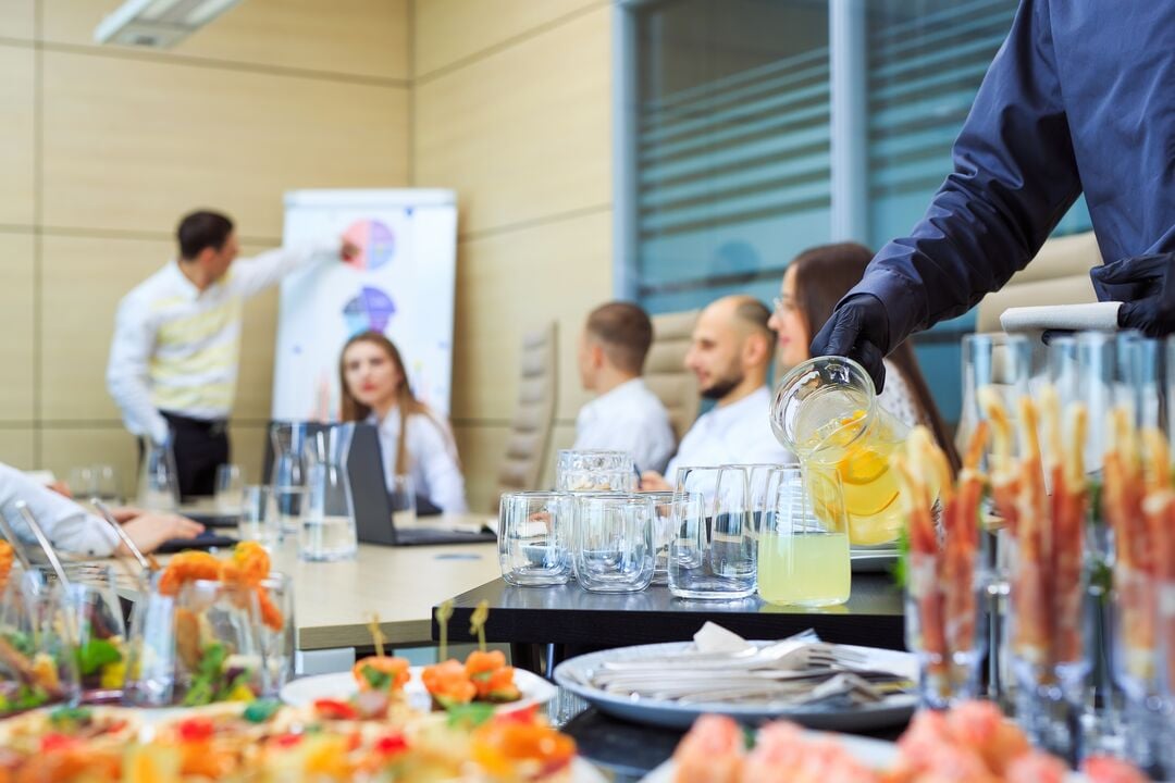 Person gießt ein Getränk aus einem Glaskrug in Trinkgläser auf einem Catering‑Tablett, im Hintergrund sitzen mehrere Personen in einem Besprechungsraum