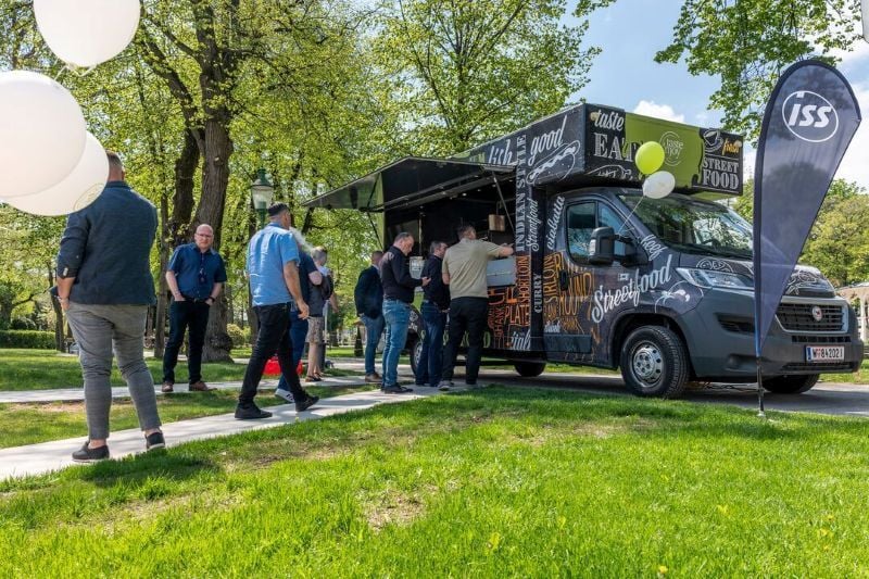 Mehrere Personen stehen im Außenbereich auf einer Grünfläche vor einem „taste ’n joy“-Street‑Food‑Truck mit geöffneter Ausgabeklappe und einer ISS‑Fahne