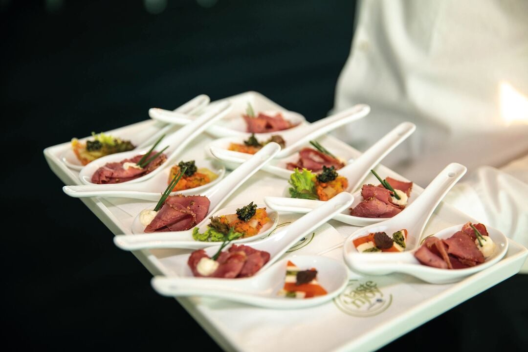 Weißes Serviertablett mit mehreren Löffeln, die mit kleinen angerichteten Häppchen aus Fleisch, Gemüse und Kräutern belegt sind, in einer Veranstaltungsumgebung