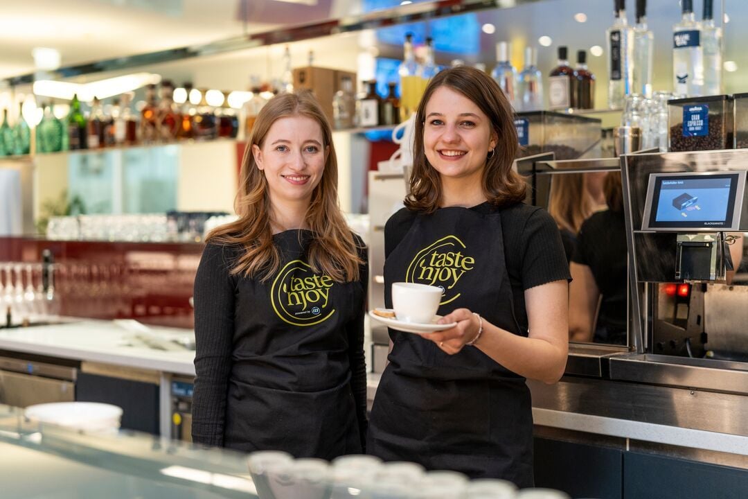 Zwei Personen mit „taste ’n joy“-Schürzen stehen hinter einem Tresen, eine Person hält eine Kaffeetasse mit Untertasse in einem gastronomischen Innenbereich