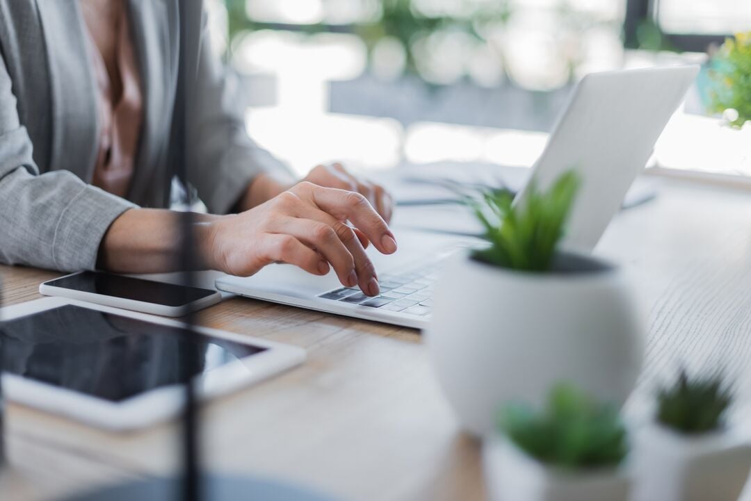 Hände einer Person tippen auf die Tastatur eines Laptops an einem Tisch mit Tablet, Smartphone und Pflanze in einem Innenbereich
