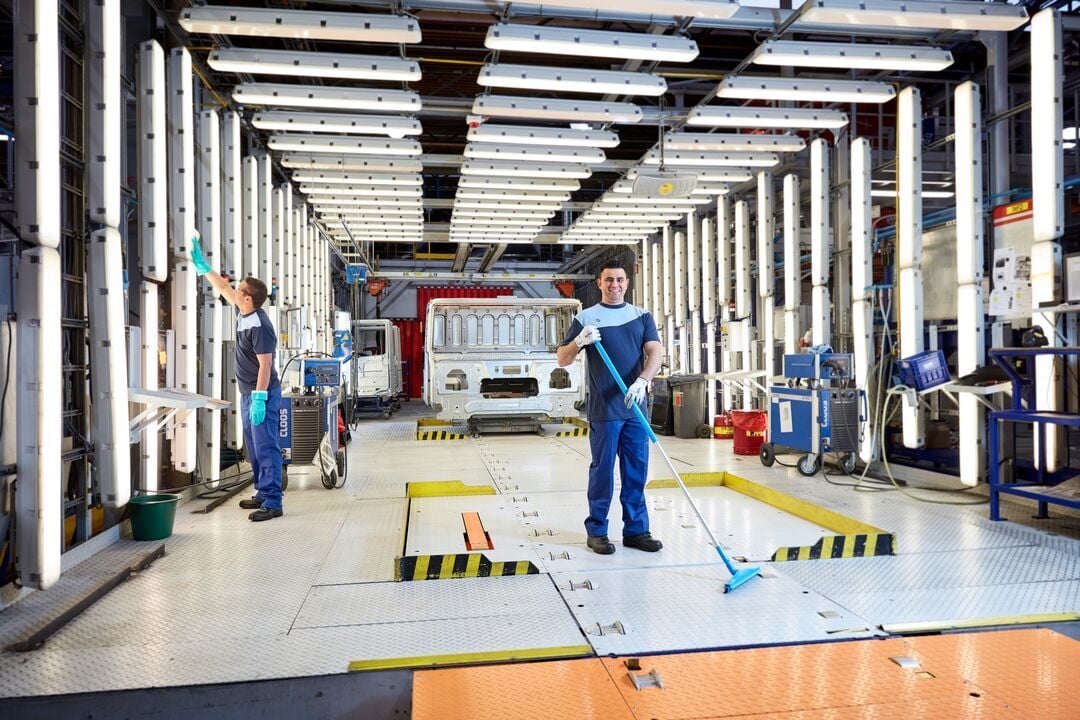 Zwei Personen in Arbeitskleidung mit Handschuhen reinigen den Boden und eine Wandfläche in einer großen Industriehalle mit technischen Anlagen