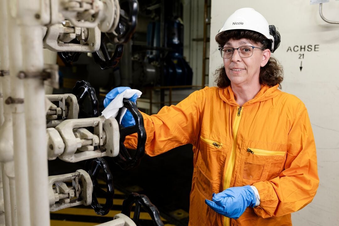 Person in Schutzkleidung mit Helm und Handschuhen wischt ein metallisches Bauteil mit Handrad in einem technischen Anlagenbereich
