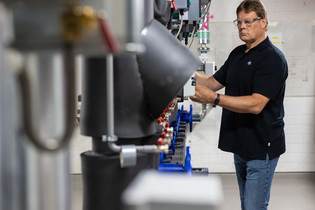 Person mit Brille bedient ein Bauteil an Rohrleitungen und Ventilen in einem technischen Anlagenraum