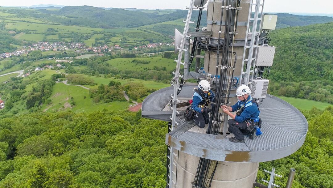 Zwei ISS Mitarbeitende in Arbeitskleidung und Schutzhelmen arbeiten gesichert auf einer runden Plattform an Kabeln und technischen Komponenten an einem hohen Bauwerk im Außenbereich