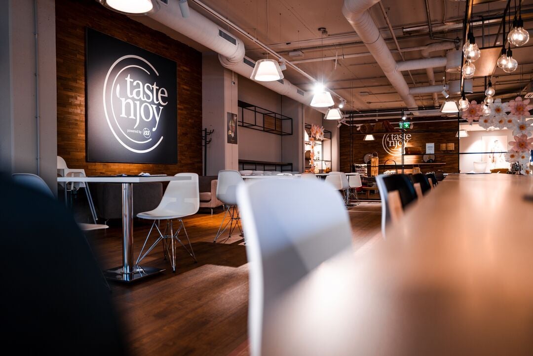 Moderner Innenbereich einer gastronomischen Einrichtung mit Tischen, Stühlen, Hängelampen und einem „taste ’n joy powered by ISS“-Logo an der Wand