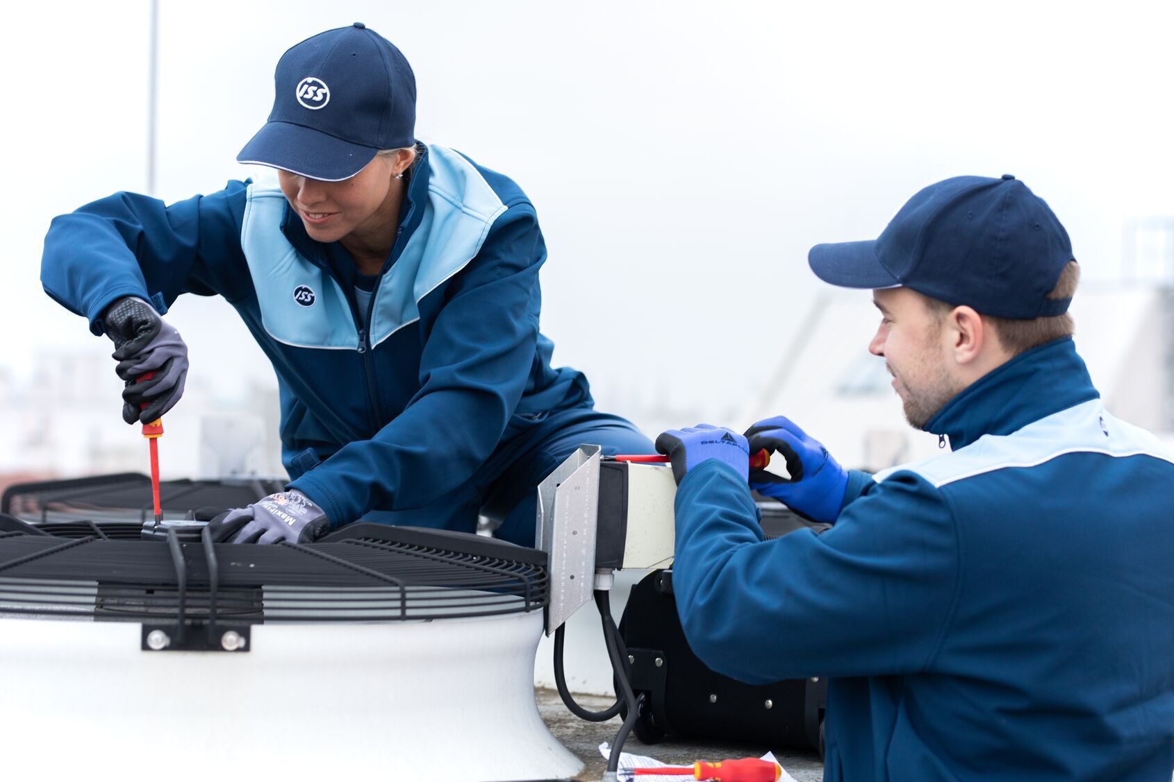 Zwei ISS Mitarbeitende in Arbeitskleidung mit Handschuhen arbeiten mit Schraubendrehern an einer technischen Anlage im Außenbereich