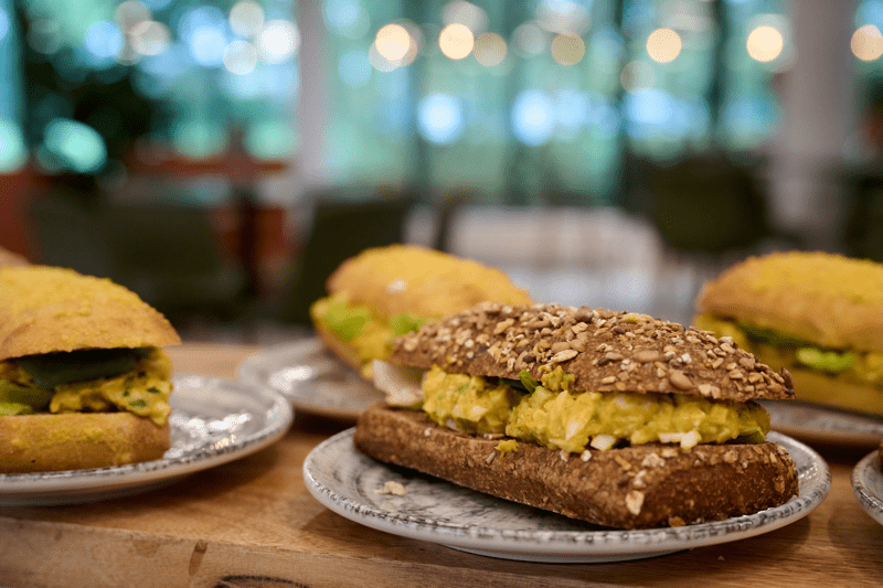 Mehrere belegte Brote auf Tellern mit körnigem und hellem Brot sowie gelber Füllung und grünen Blättern auf einer Holzoberfläche