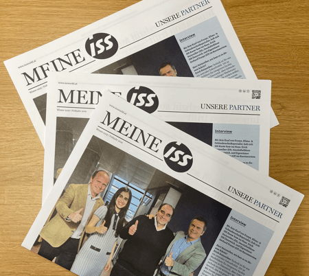 Mehrere Exemplare der Zeitung ‚Meine ISS‘ liegen leicht überlappend auf einer Holzoberfläche, auf der Titelseite ist eine Gruppe von Personen abgebildet.