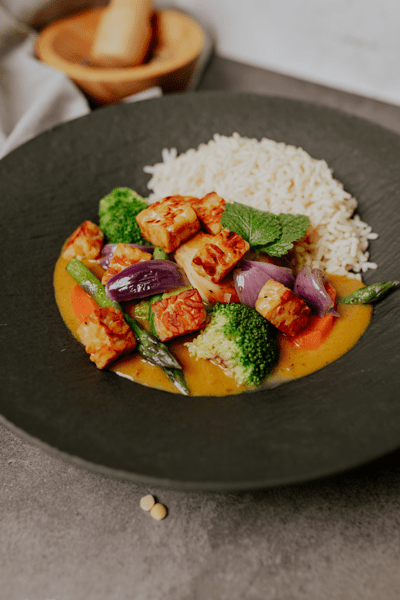 Teller mit Reis und gebratenen Würfeln aus pflanzlichem Protein in gelber Sauce, serviert mit Brokkoli, Karotten, Spargel und roten Zwiebeln.