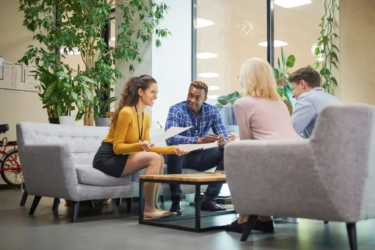Vier Mitarbeitende besprechen Unterlagen in einer modernen Büro Lounge mit Pflanzen und lockerer Meeting Atmosphäre zur Förderung von Zusammenarbeit